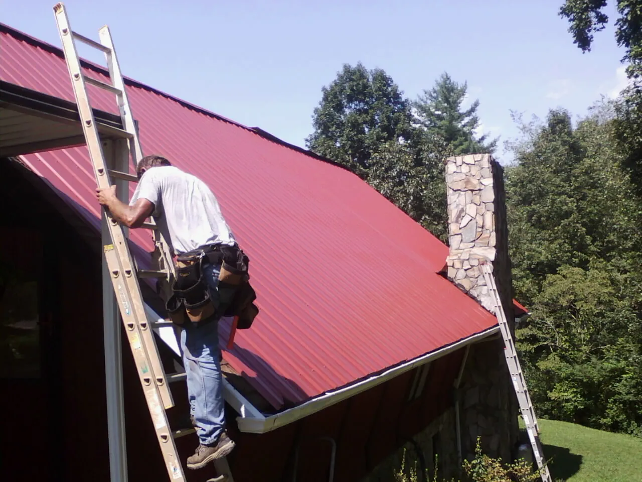 Roofer on a ladder beside a red metal roof and stone chimney — Soffit & Fascia Repair by Roofing Brothers in Pawtucket